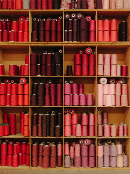 Shelved spools of red, burgundy, and pink thread organized in wooden compartments.