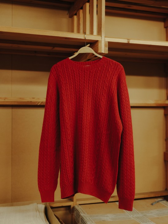 Red cable-knit cashmere jumper hanging on a wooden hanger against a pale yellow wall.