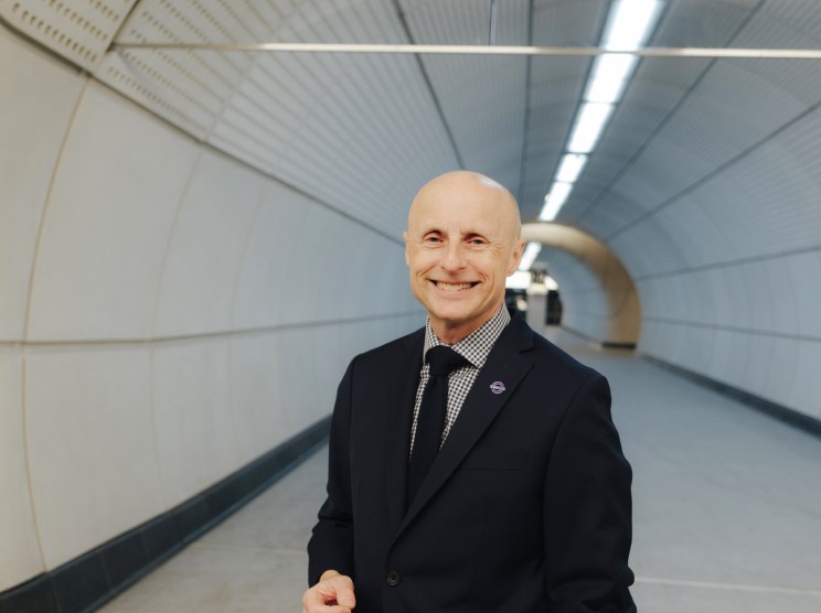 Andy Byford in a dark suit standing in a modern underground tunnel with curved white walls and fluorescent lighting