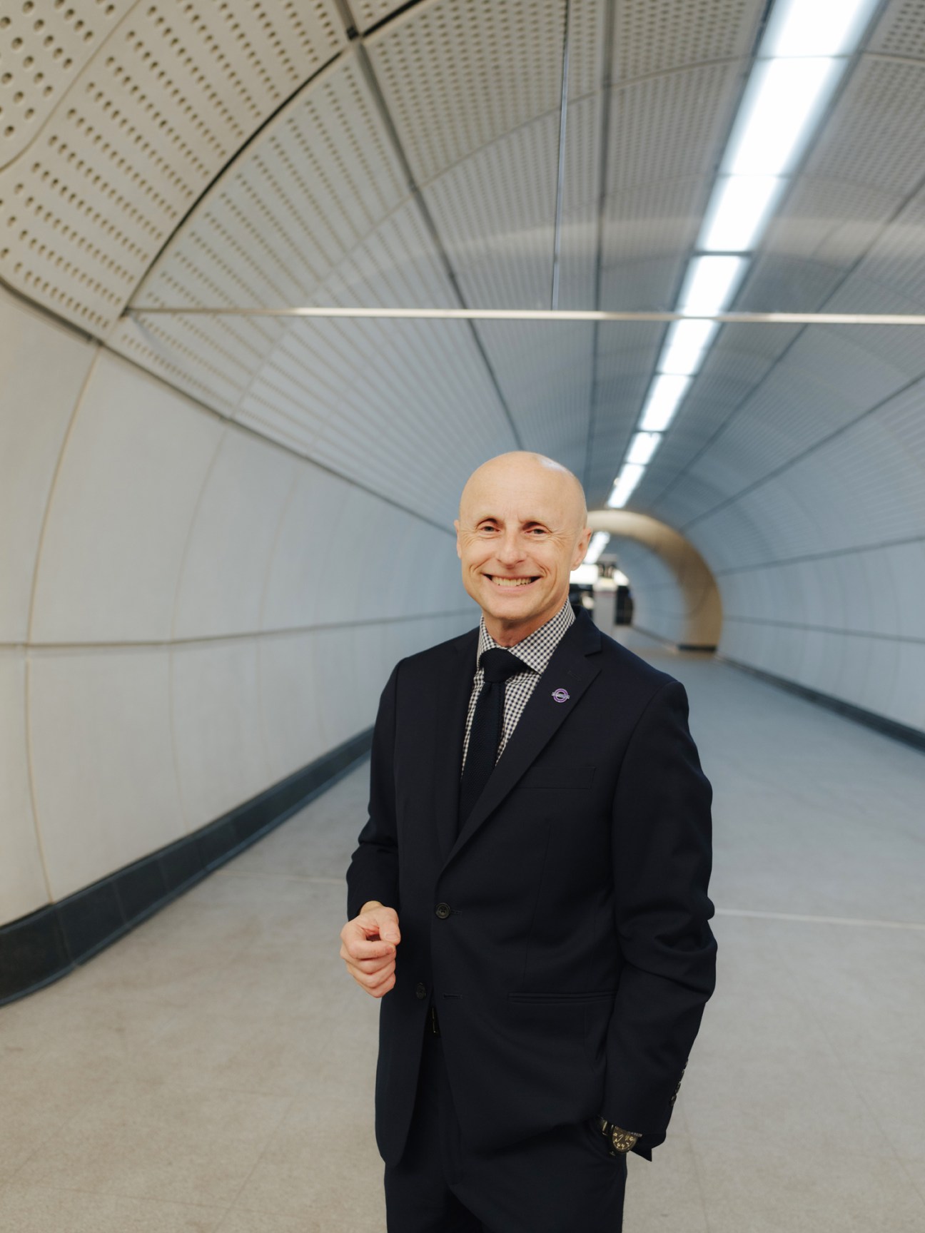 Andy Byford in a dark suit standing in a modern underground tunnel with curved white walls and fluorescent lighting