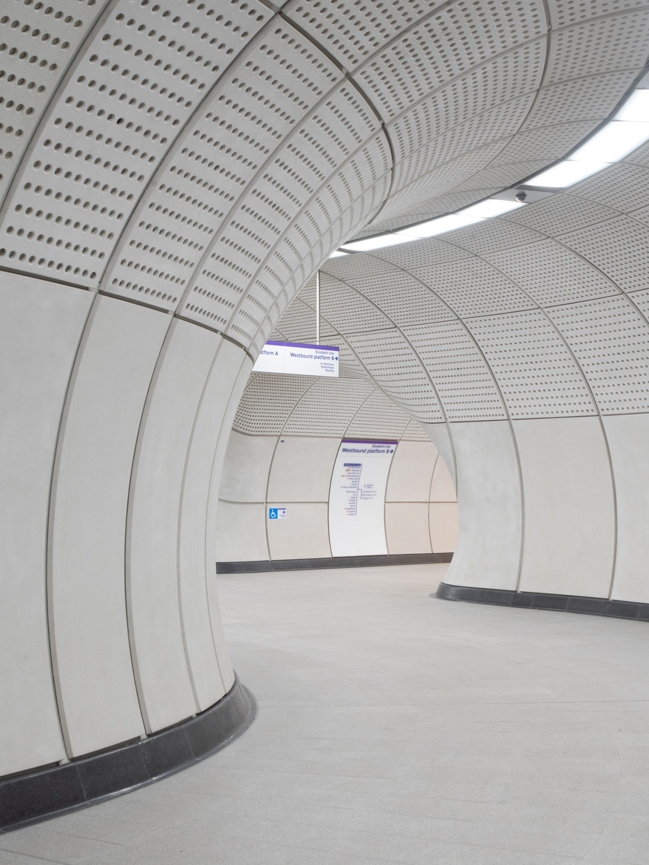Modern underground railway platform with curved white walls, perforated ceiling panels, and platform benches