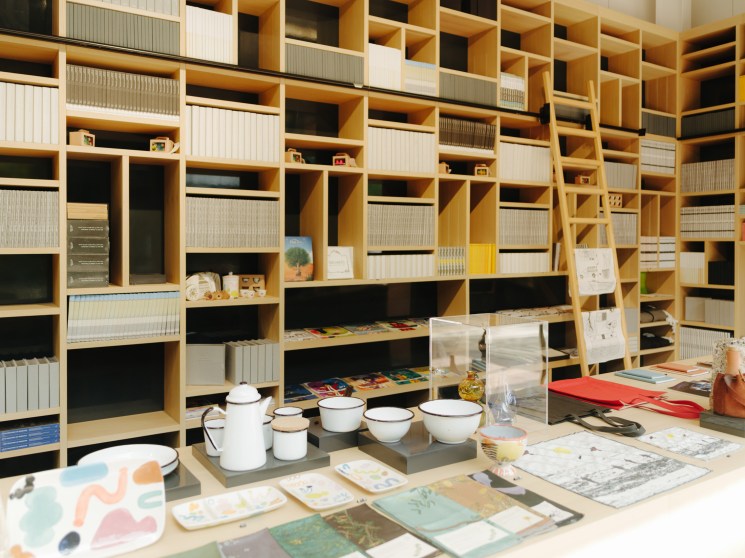 Interior of a modern retail space with wooden shelving, display items including ceramics and books, and design materials on a table.