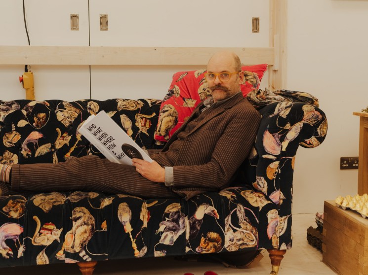 A bearded man in a suit reclines on a bold floral-patterned sofa reading a magazine, shoes discarded on the floor beside him.