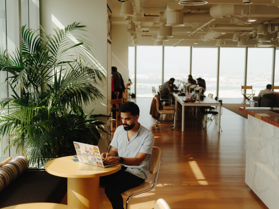 Man working on laptop at a table in a modern co-working space with large windows and palm plants.