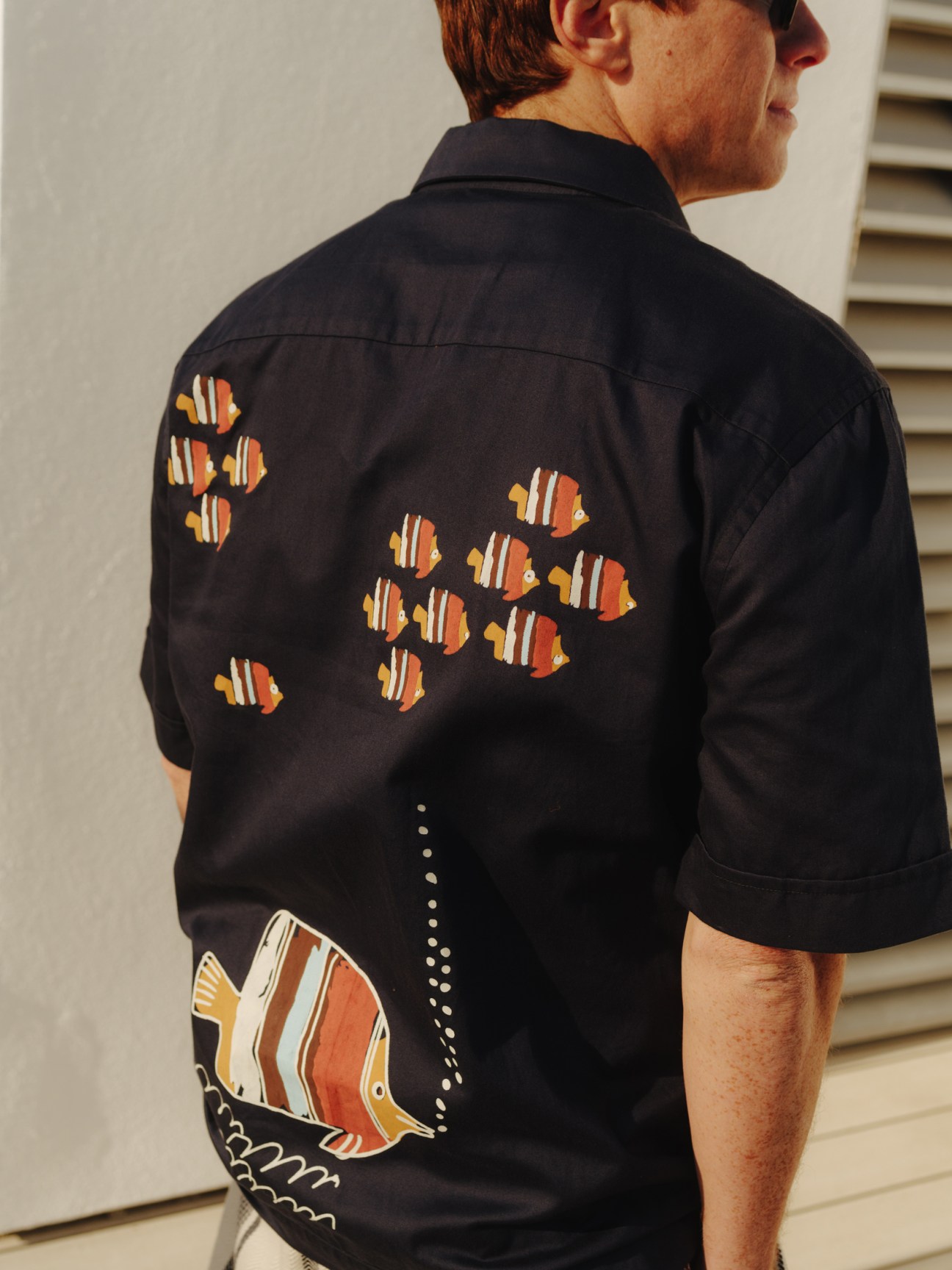 Man wearing black short-sleeved shirt with embroidered colorful fish motifs on back, viewed from behind.