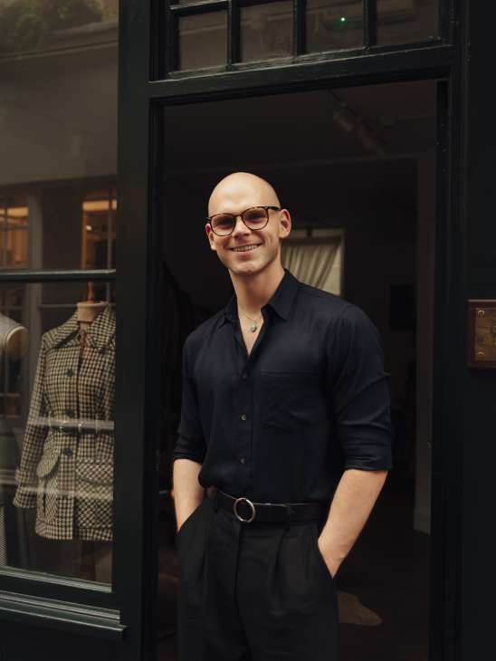 Joshua Millard standing in doorway of his studio-shop wearing black shirt and glasses
