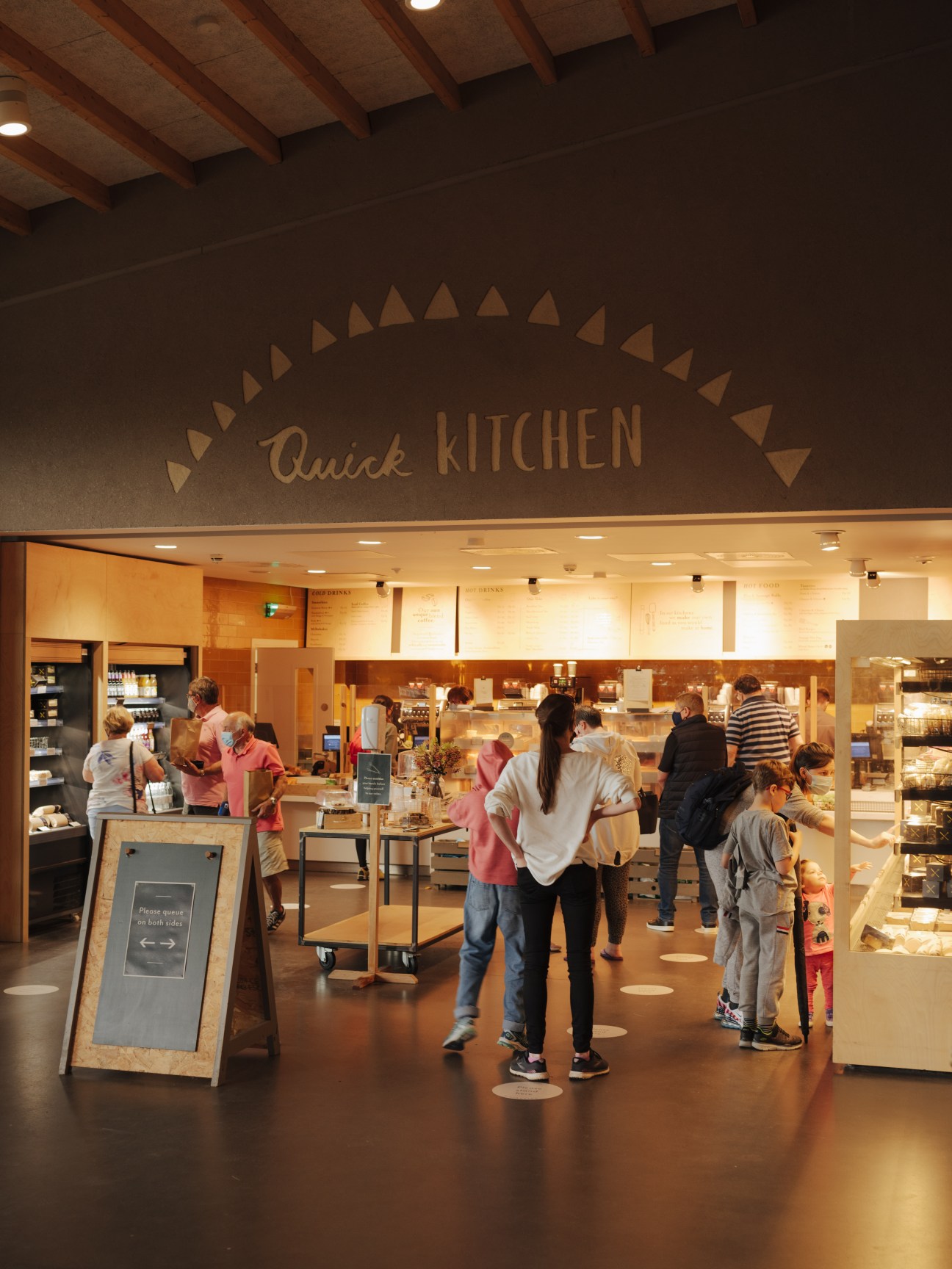 Customers queue at a Quick Kitchen counter inside Gloucester Services with warm wood and lighting.