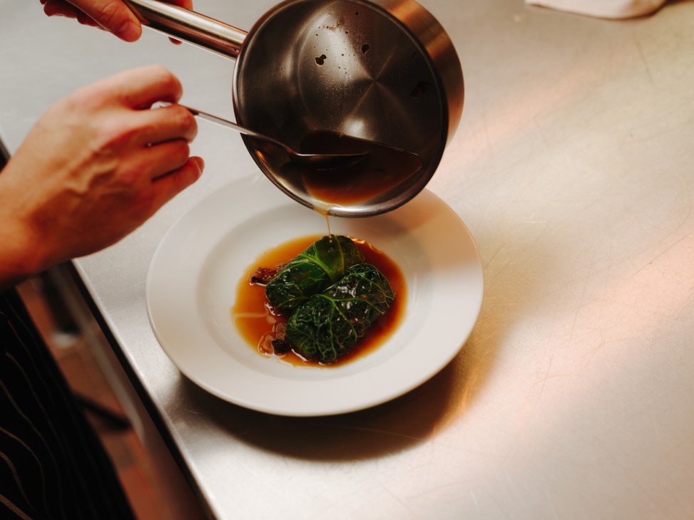 Chef pouring dark sauce from a saucepan onto a plate with leafy greens in restaurant kitchen