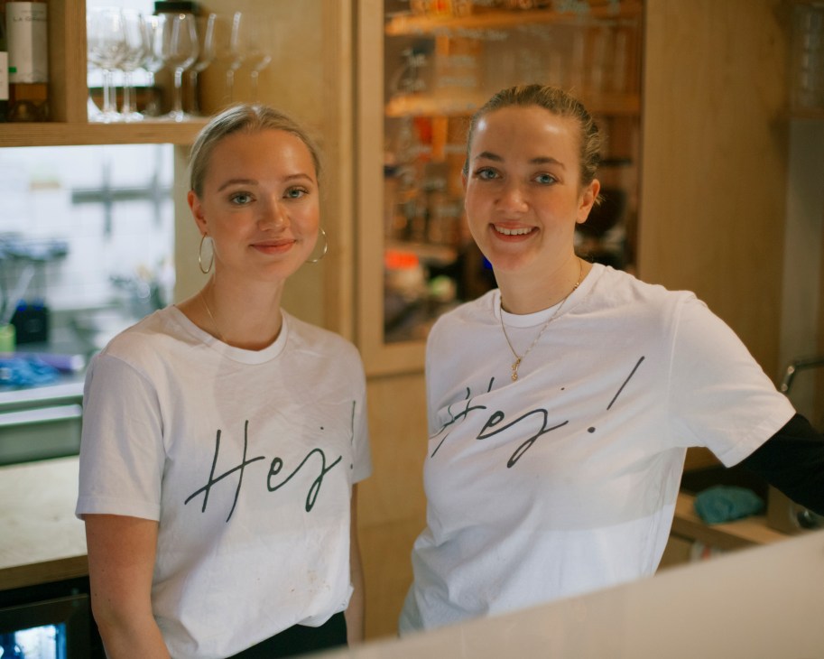 Two women in matching white t-shirts with 'Hej' written in script, standing in a Nordic-style interior