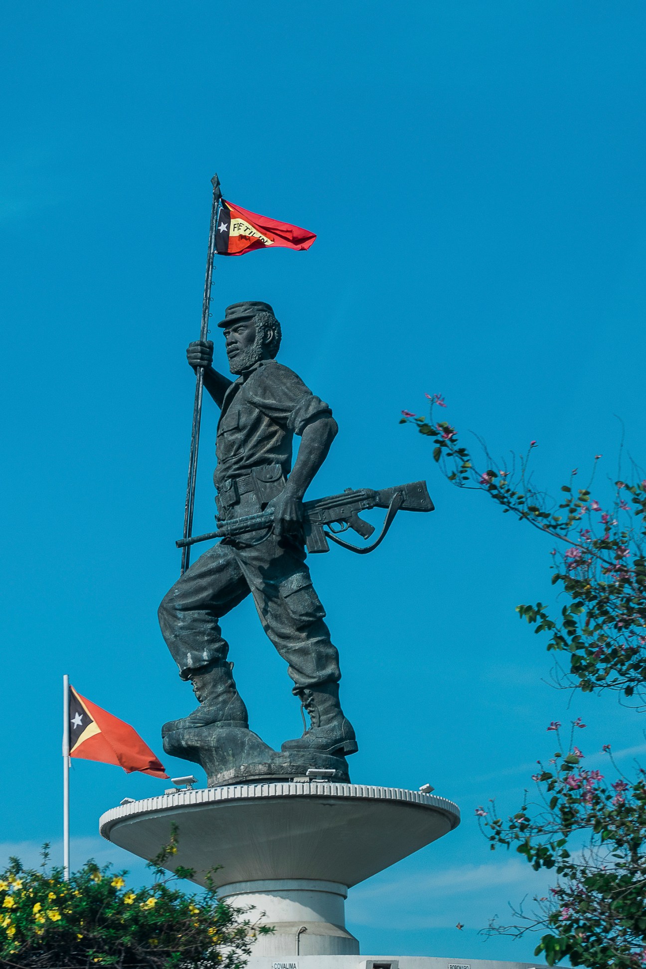 Bronze statue of armed soldier holding flag on pedestal with East Timor flag visible in background