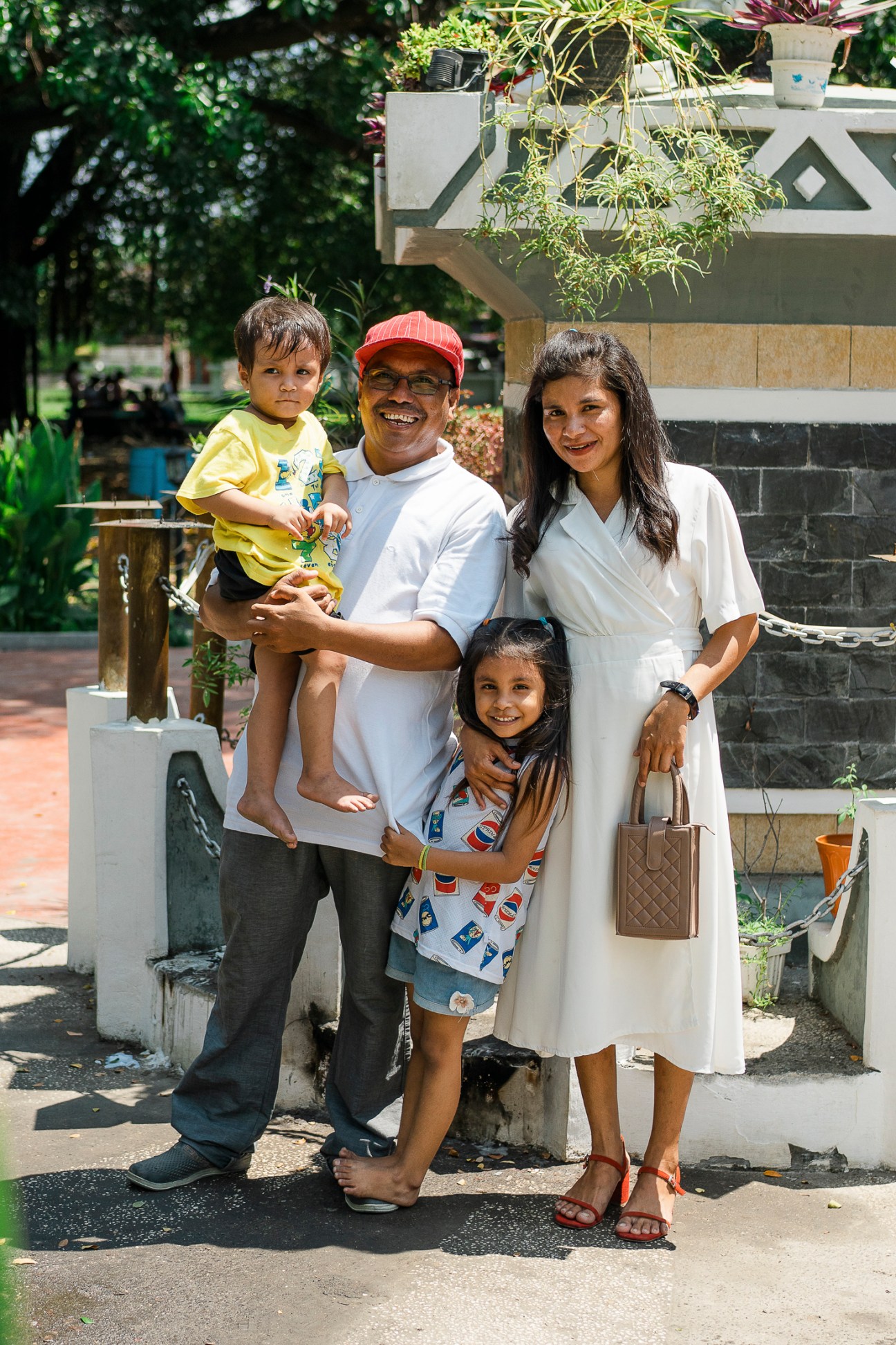 A happy family of four poses together outdoors in Dili - parents and two young children smiling at the camera