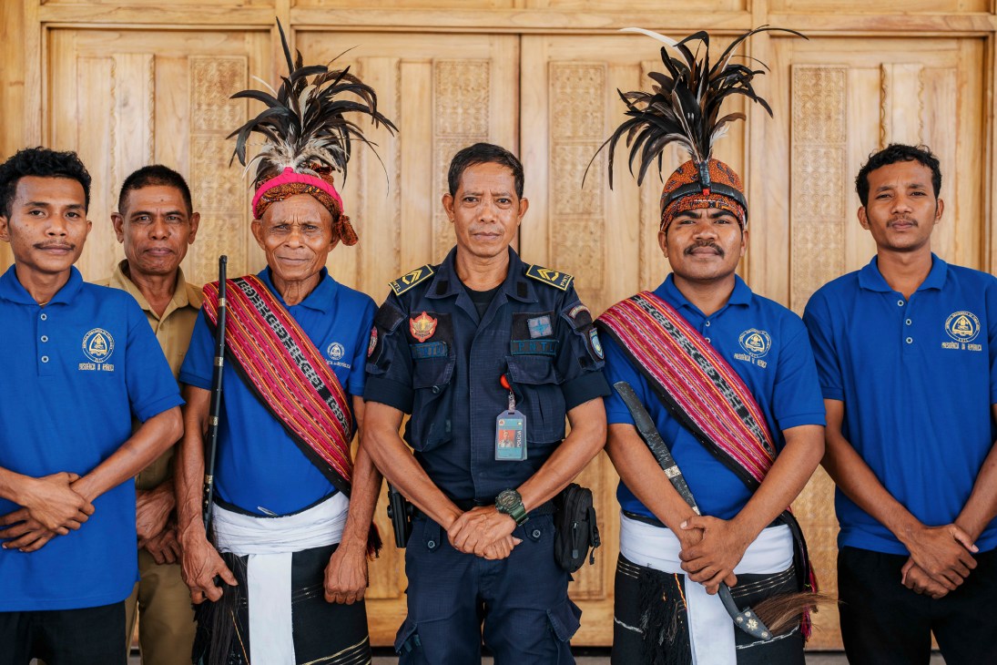 Six men in formal attire stand in a line, two wearing traditional feathered headdresses and ceremonial sashes