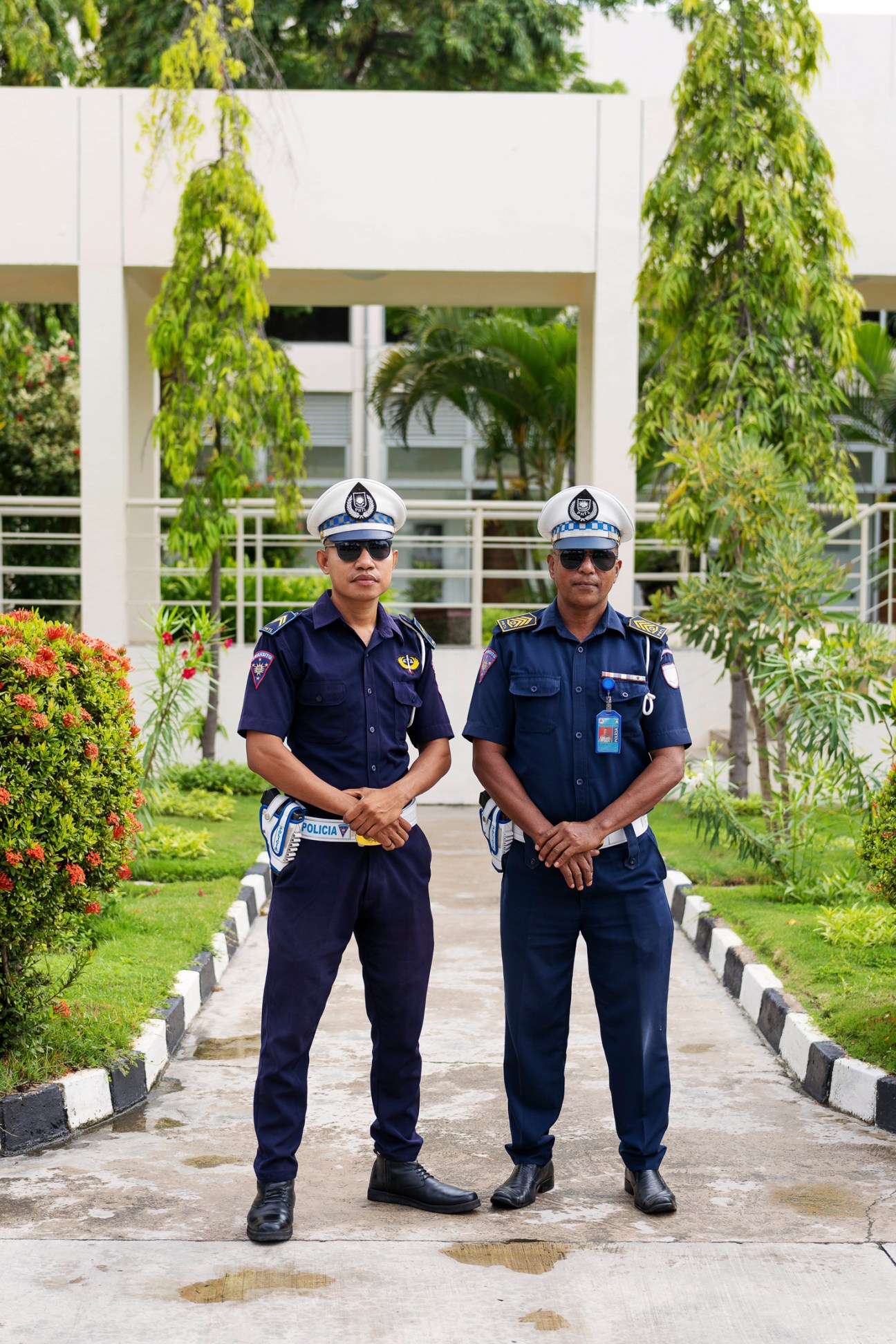 Two uniformed traffic police officers in white caps and dark blue uniforms stand on a paved walkway in Dili