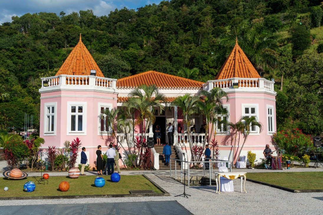 Pink colonial palace with orange tile roofs and twin towers, people gathering on front steps, tropical setting