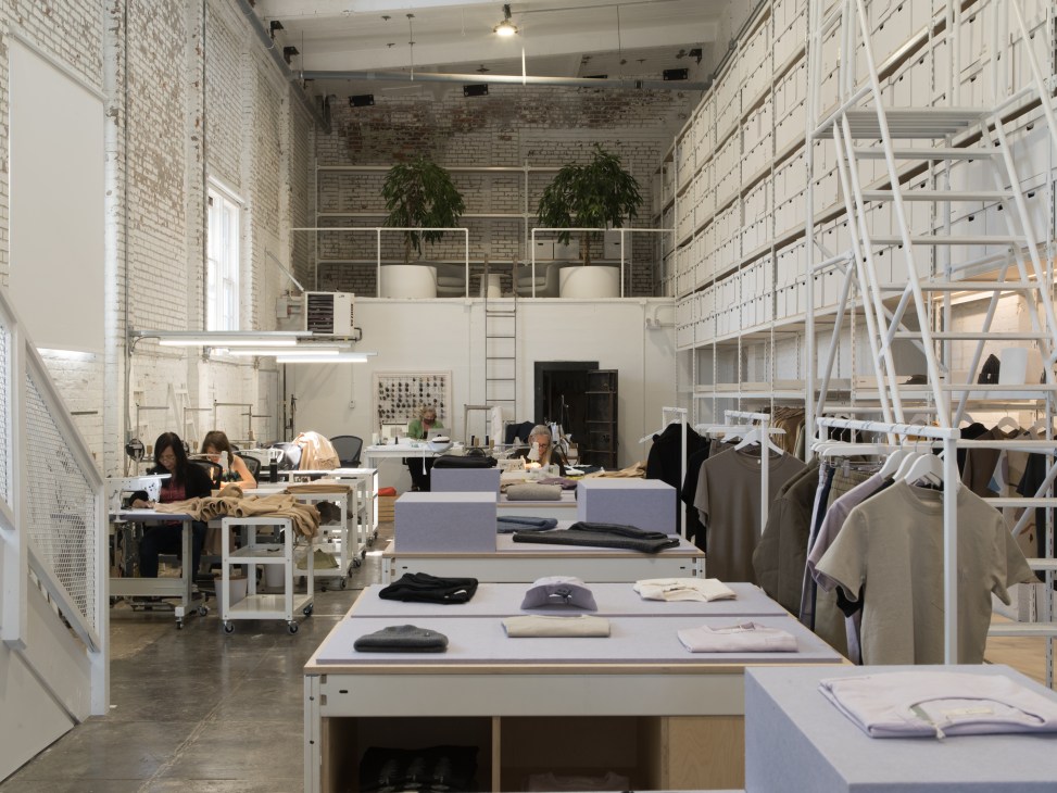 Interior of Ecologyst's manufacturing facility showing sewing stations with workers and white shelving units stacked with inventory boxes.