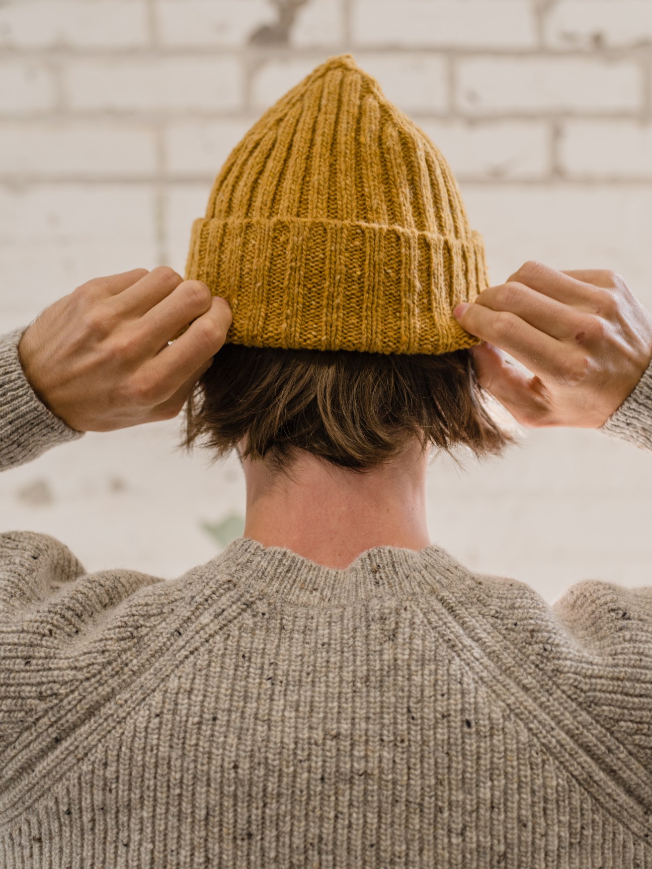 Person wearing a mustard yellow knit beanie and gray ribbed sweater against a white brick wall.