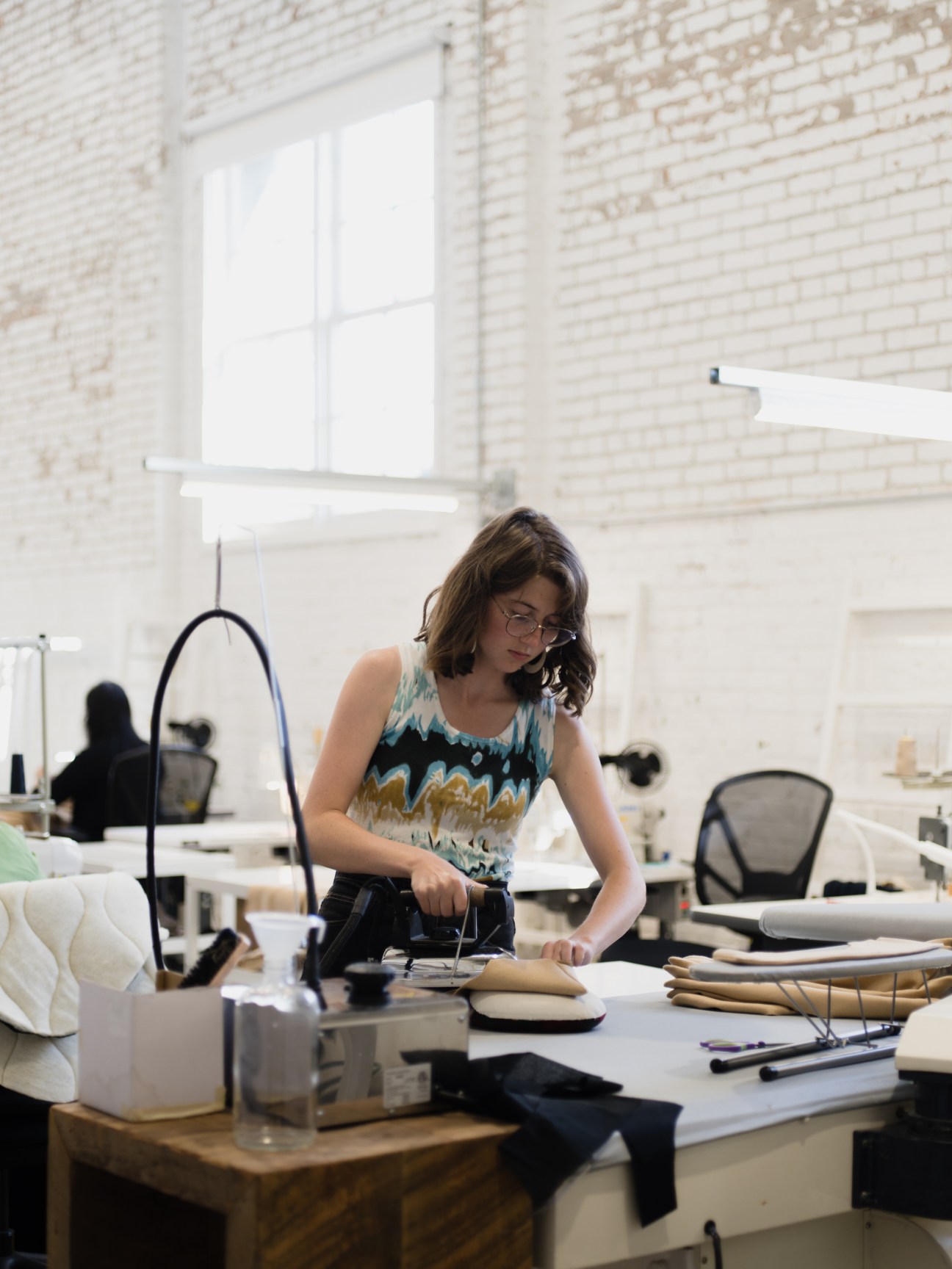 Woman working at a sewing table in a bright studio space with white brick walls and large windows.