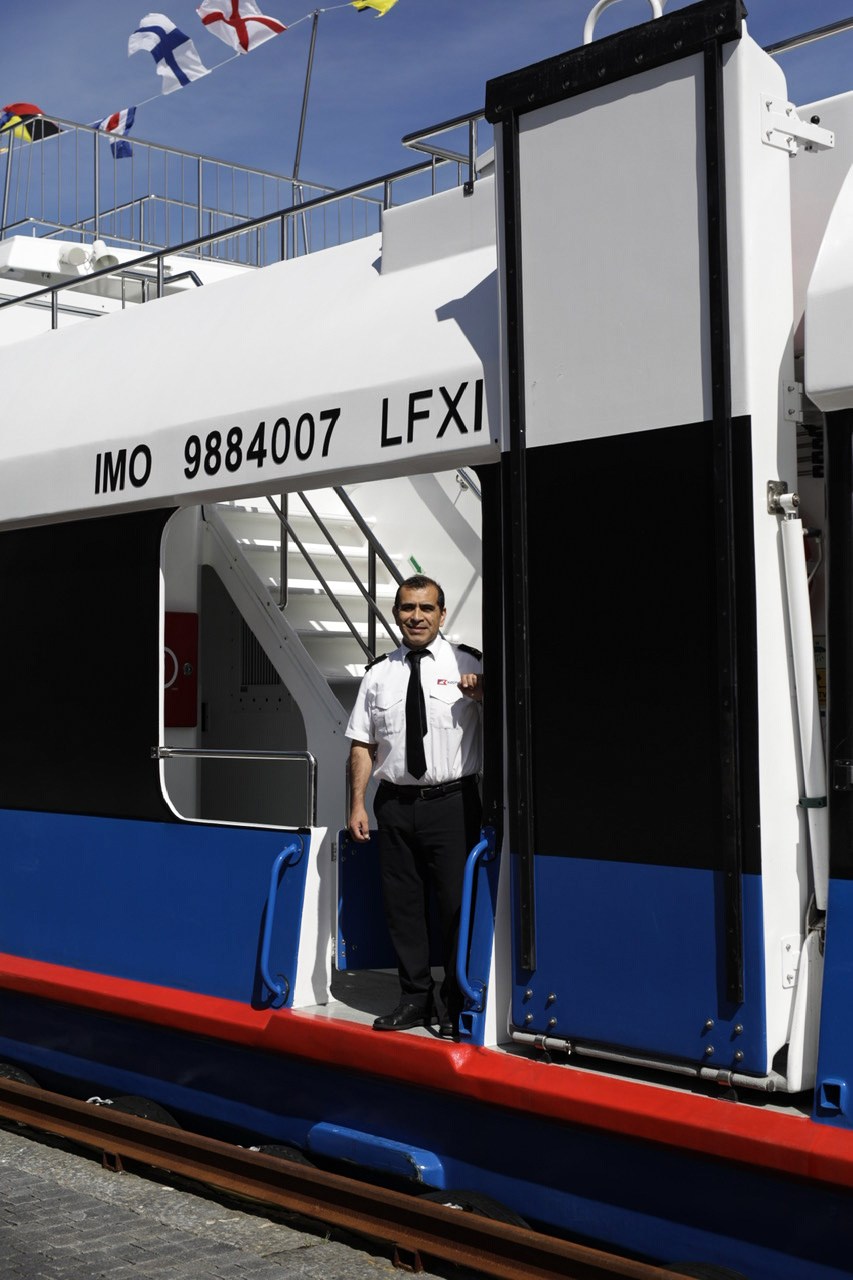 Ship crew member in white uniform standing in doorway of vessel IMO 9884007 LFXI with maritime flags above