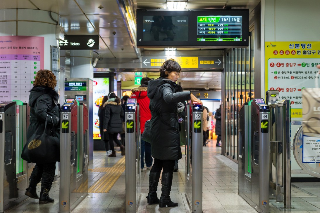 Commuters passing through ticket gates at a Seoul subway station with digital displays and signage in Korean.
