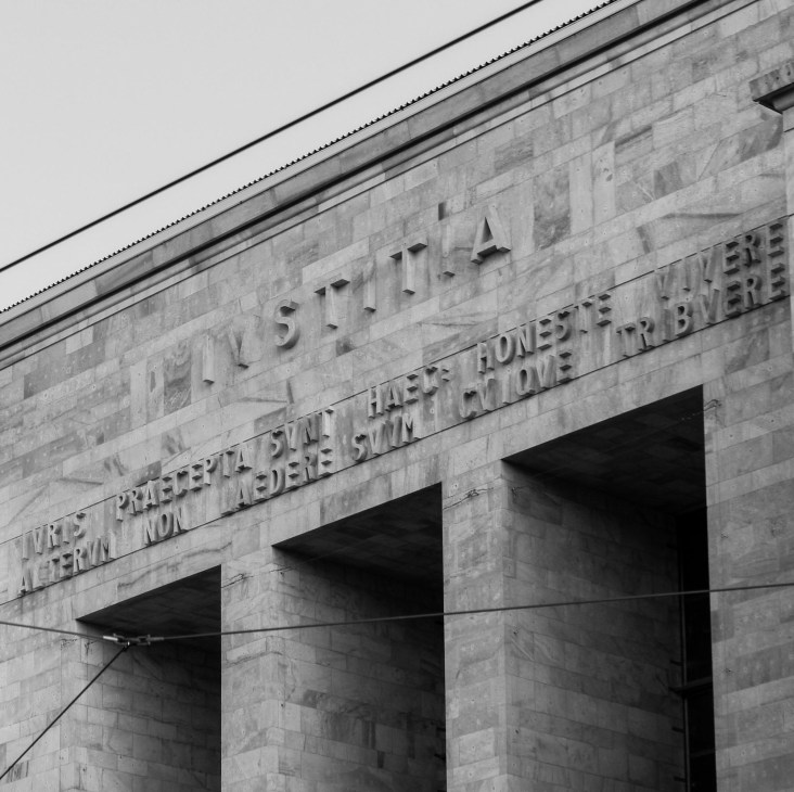 Black and white photograph of a modernist stone building facade with carved Latin text and three arched openings.
