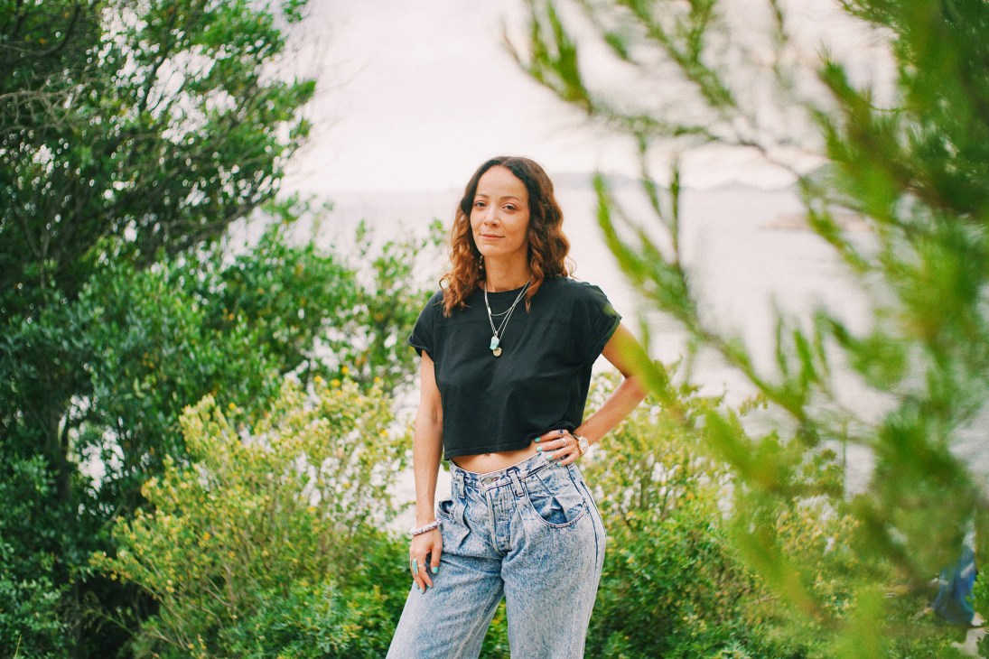 Élodie Rama stands outdoors in casual clothing with lush green foliage in the background