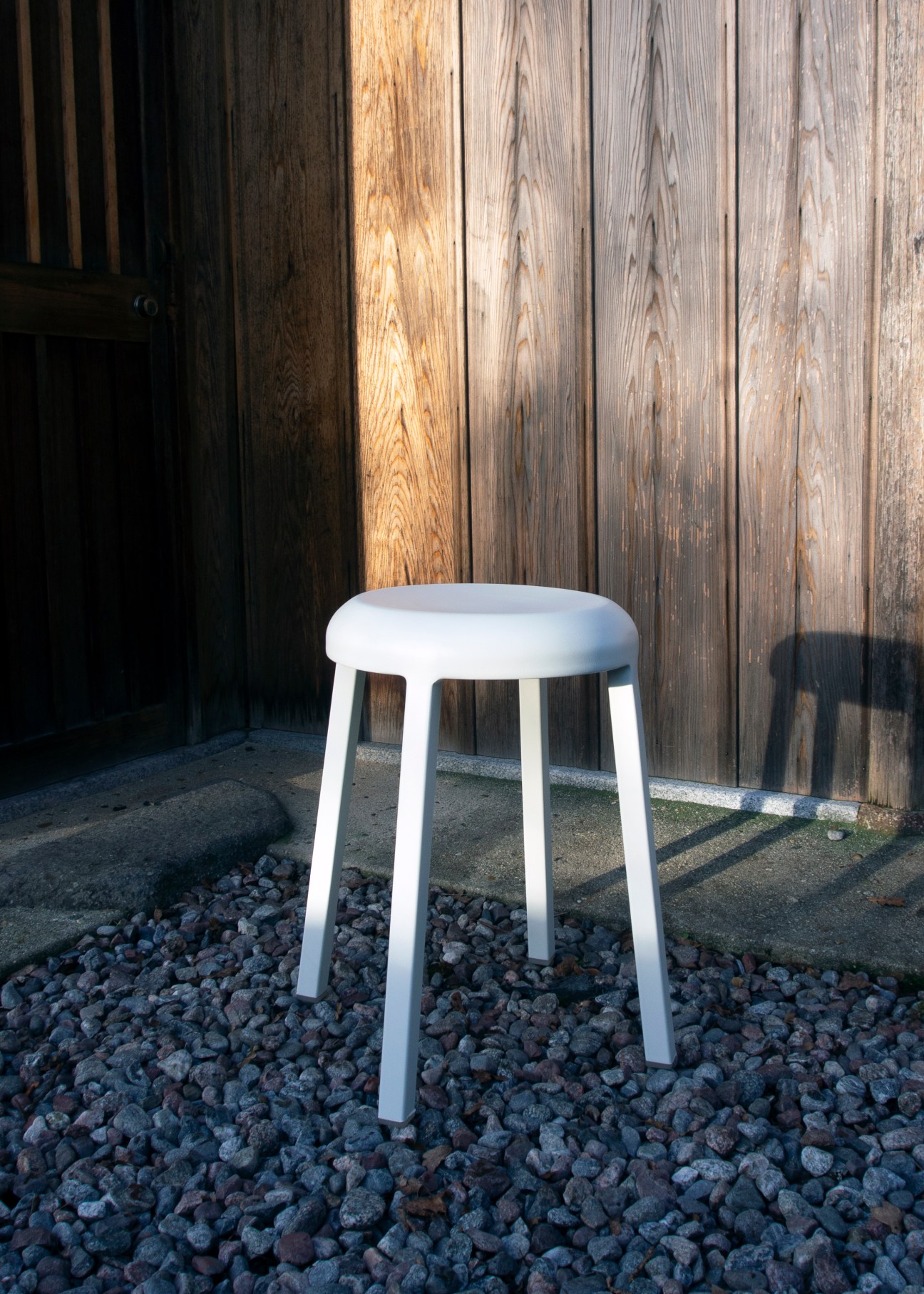 White Emeco Za recycled aluminum stool on gravel ground against weathered wooden fence in natural lighting