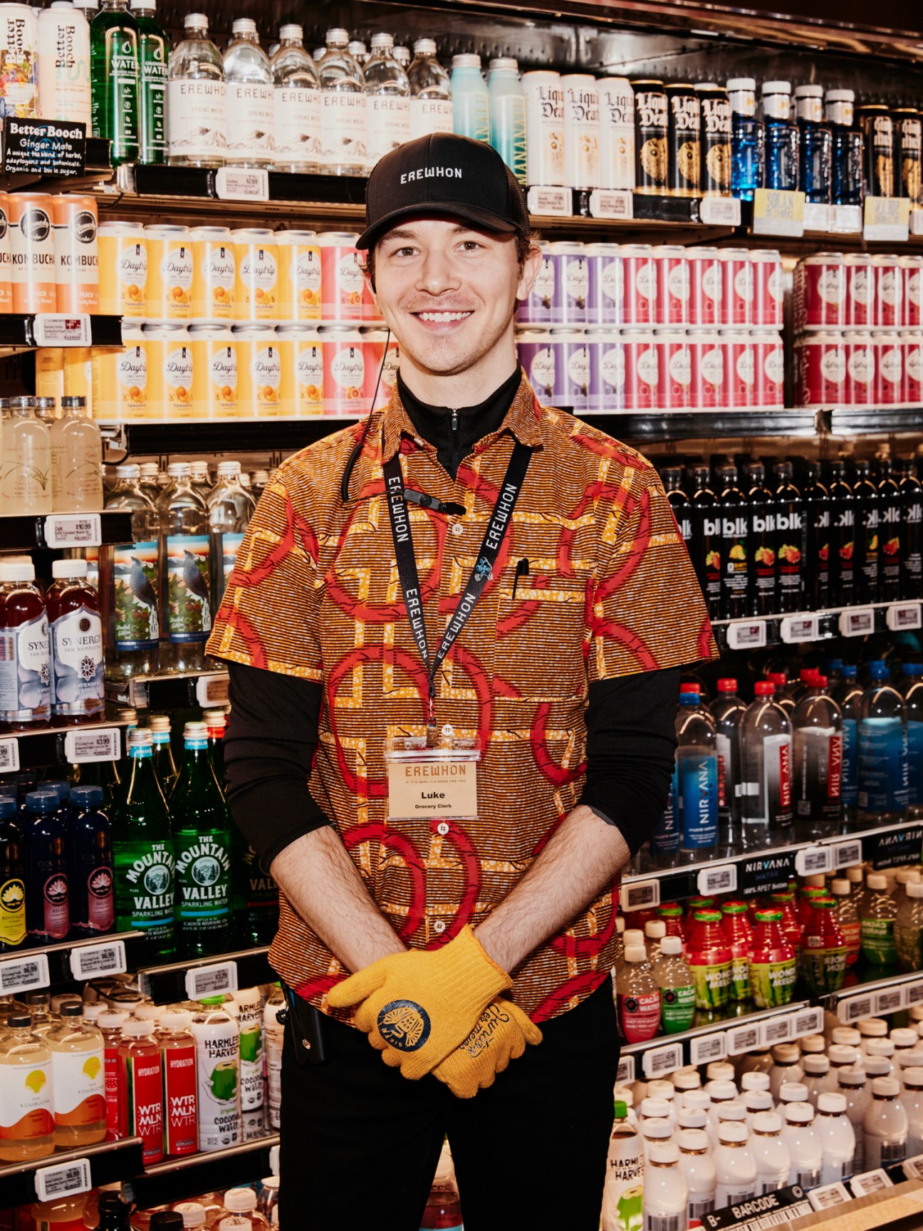 Erewhon employee wearing cap and patterned shirt stands smiling among grocery store shelves stocked with health products