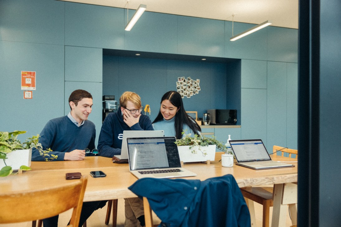 Three students collaborate at a wooden table with laptops in a modern office space with blue walls.
