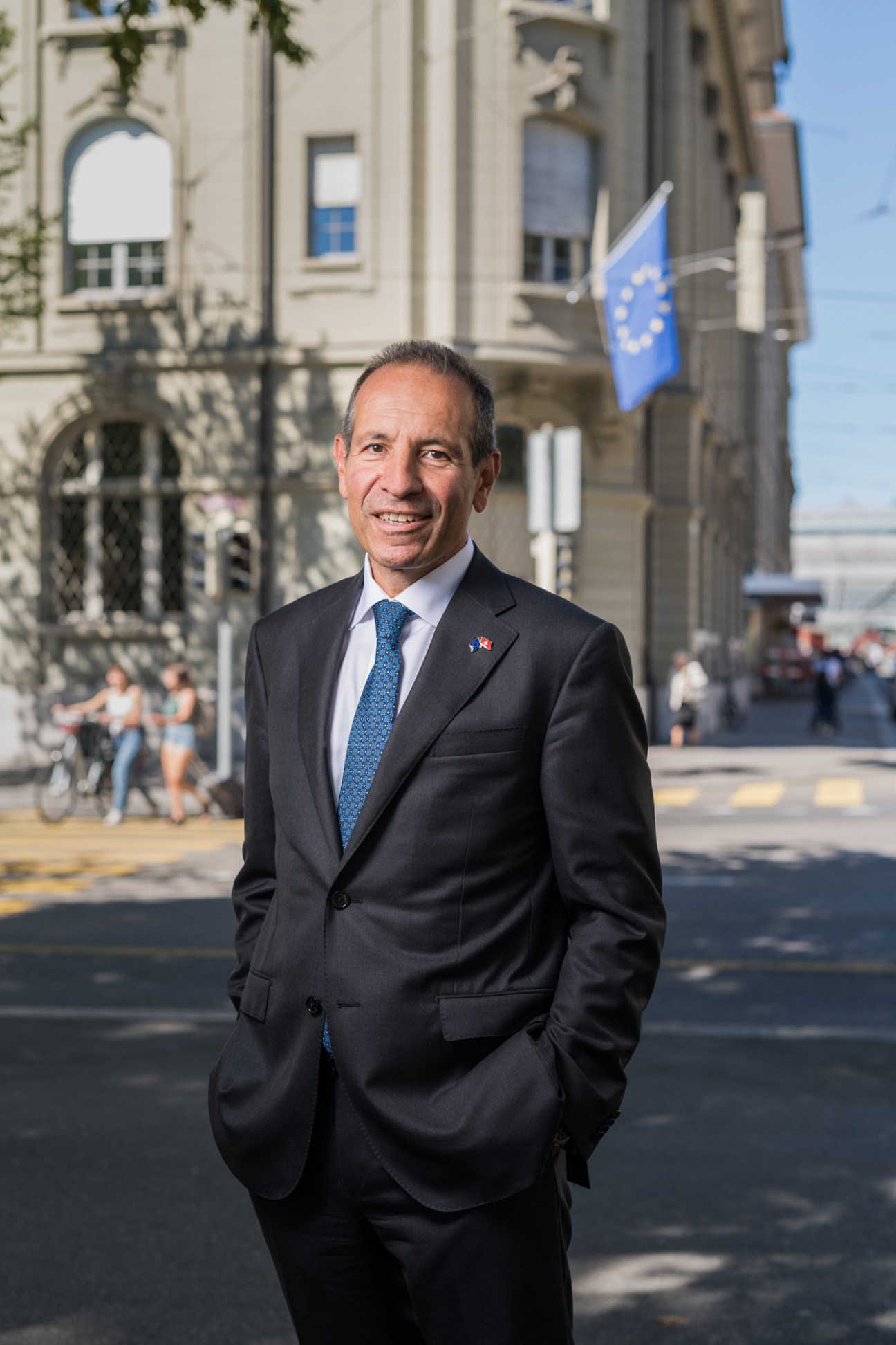 Petros Mavromichalis in a dark suit and blue tie standing on a street with European buildings in background