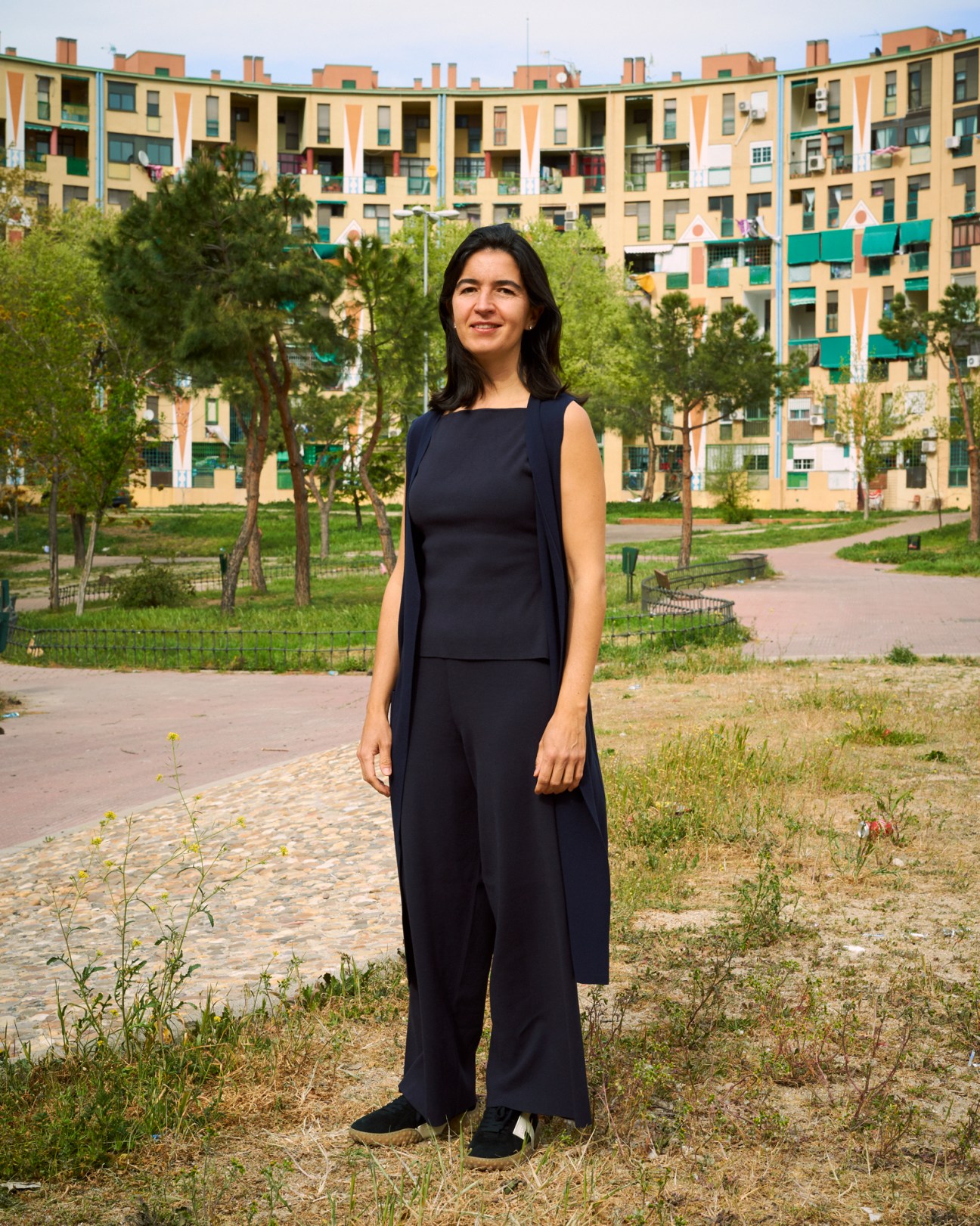 Woman in dark outfit standing in park area with colorful apartment buildings in background