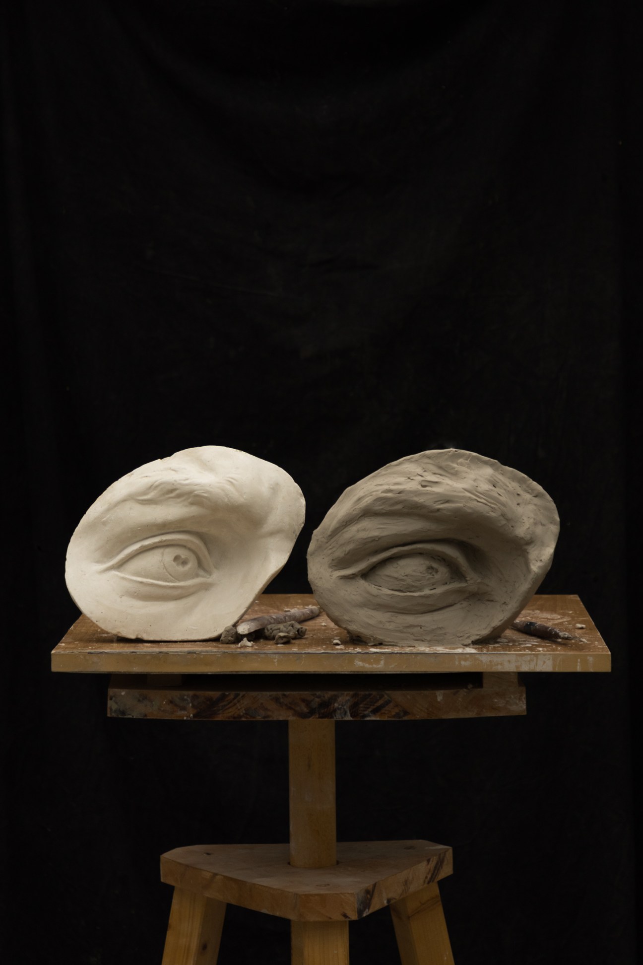 Two clay sculptures of eyes on a wooden work surface in a sculpture workshop