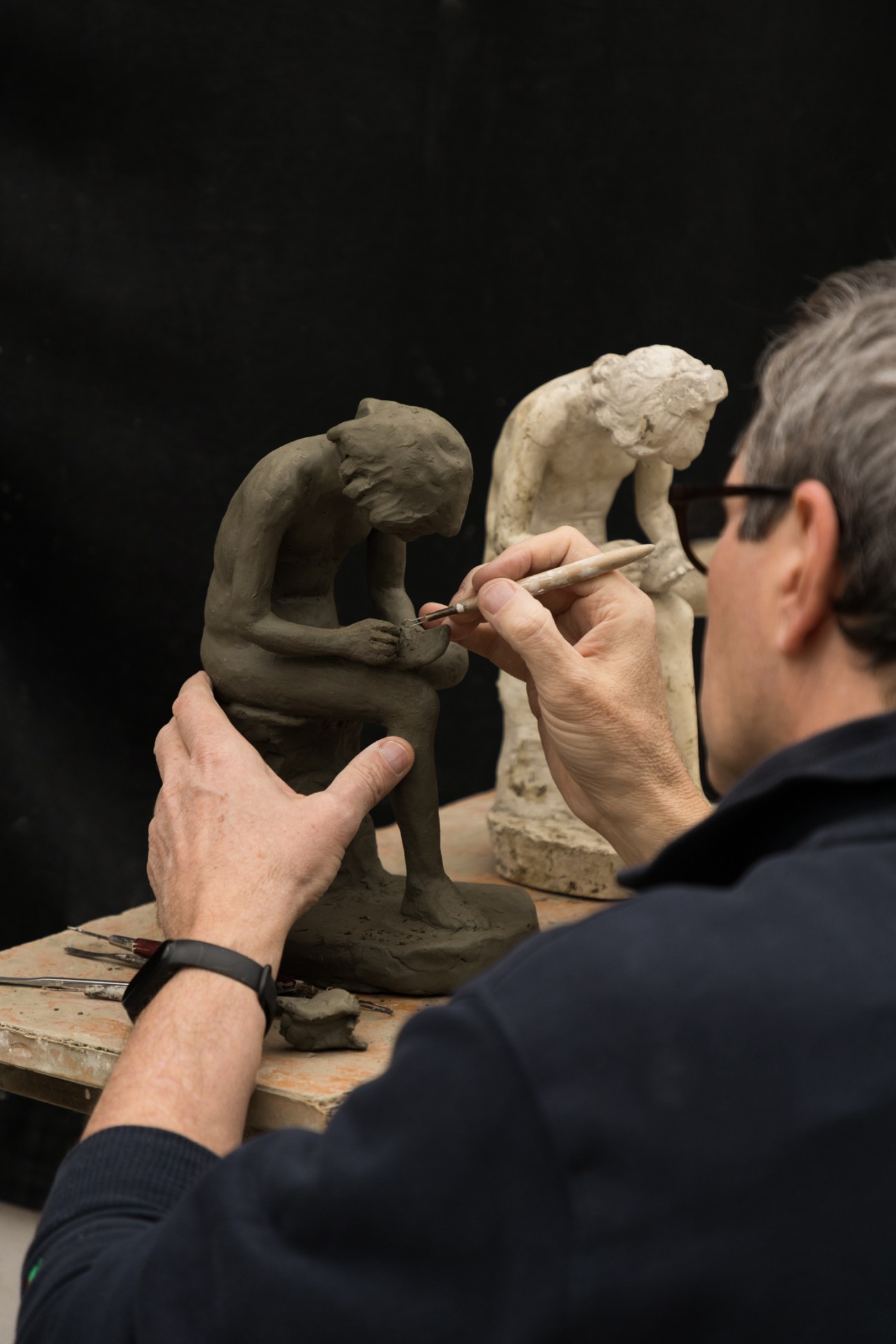 A sculptor works on a clay figure while using a plaster reference sculpture in a workshop