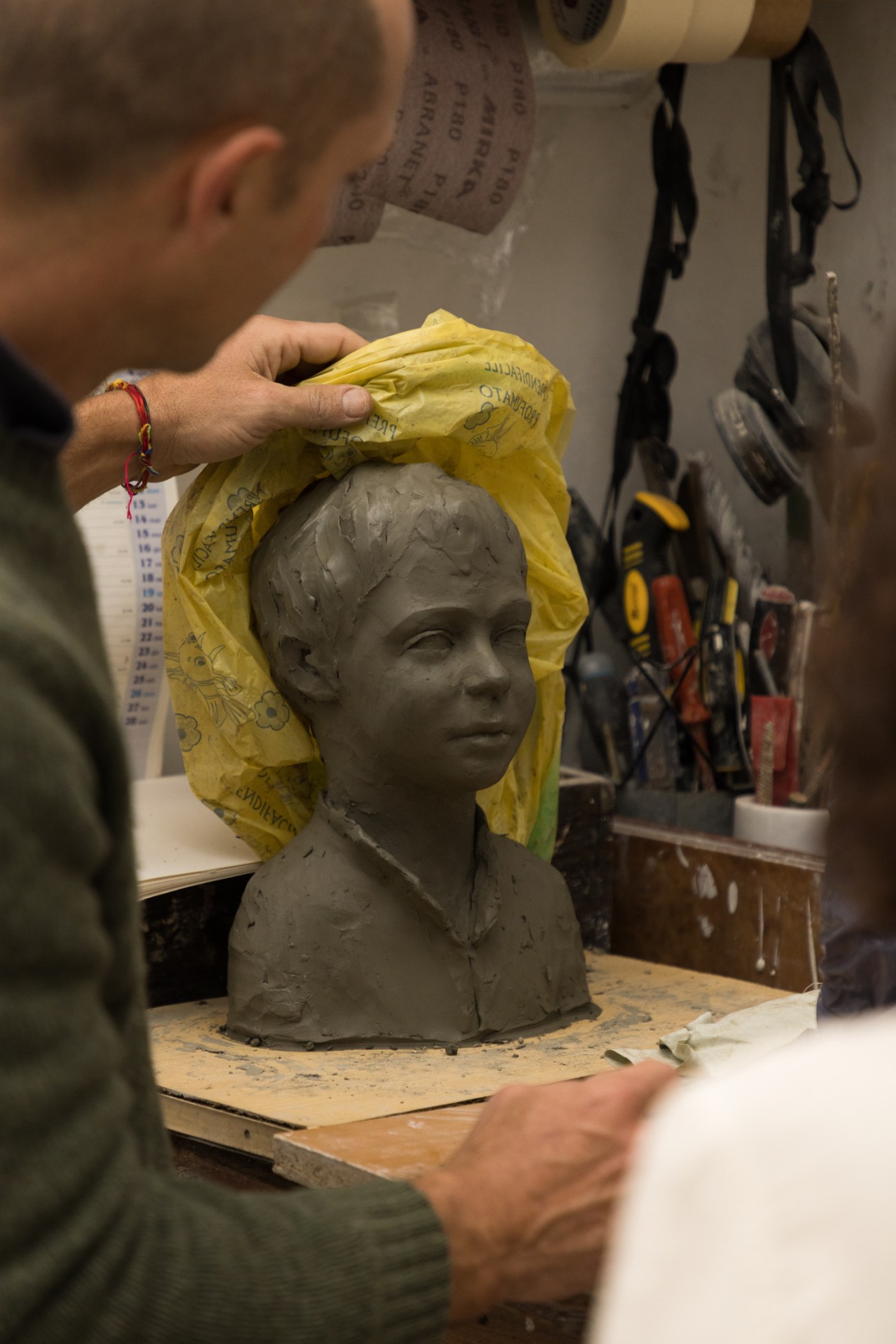 A person carefully removes a yellow plastic bag from around a clay sculpture bust in a workshop setting
