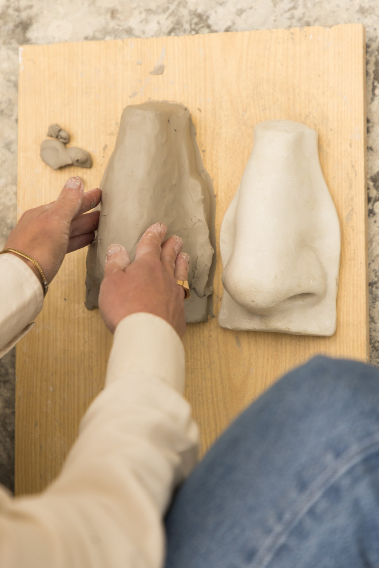 Hands sculpting clay nose on wooden board next to white plaster model in sculpture workshop