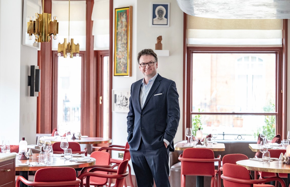 Ewan Venters stands in an elegant restaurant dining room with red chairs and brass fixtures.