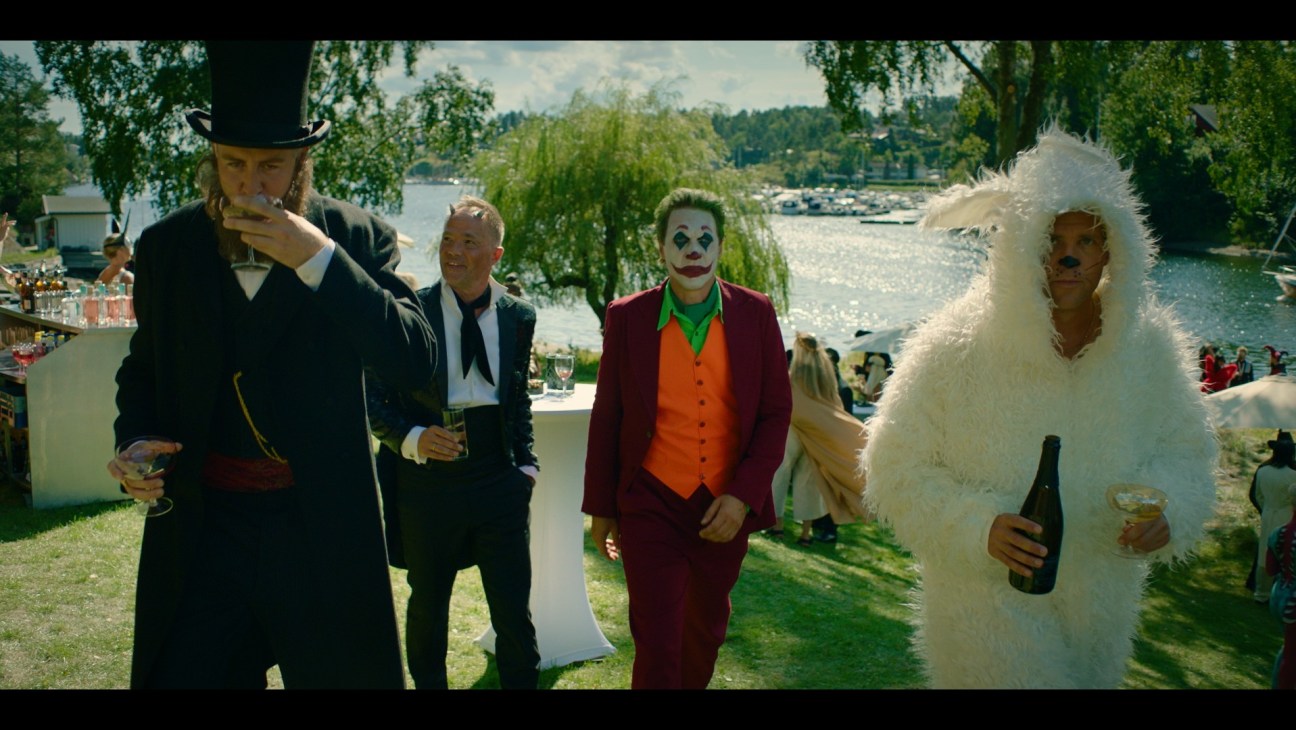 Four men in costume at outdoor event by waterfront - one in naval uniform, one in suit, one as Joker, one in white fur coat