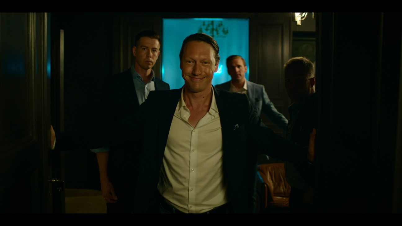 Man in dark jacket and white shirt smiling while walking with other men in suits behind him in dimly lit corridor