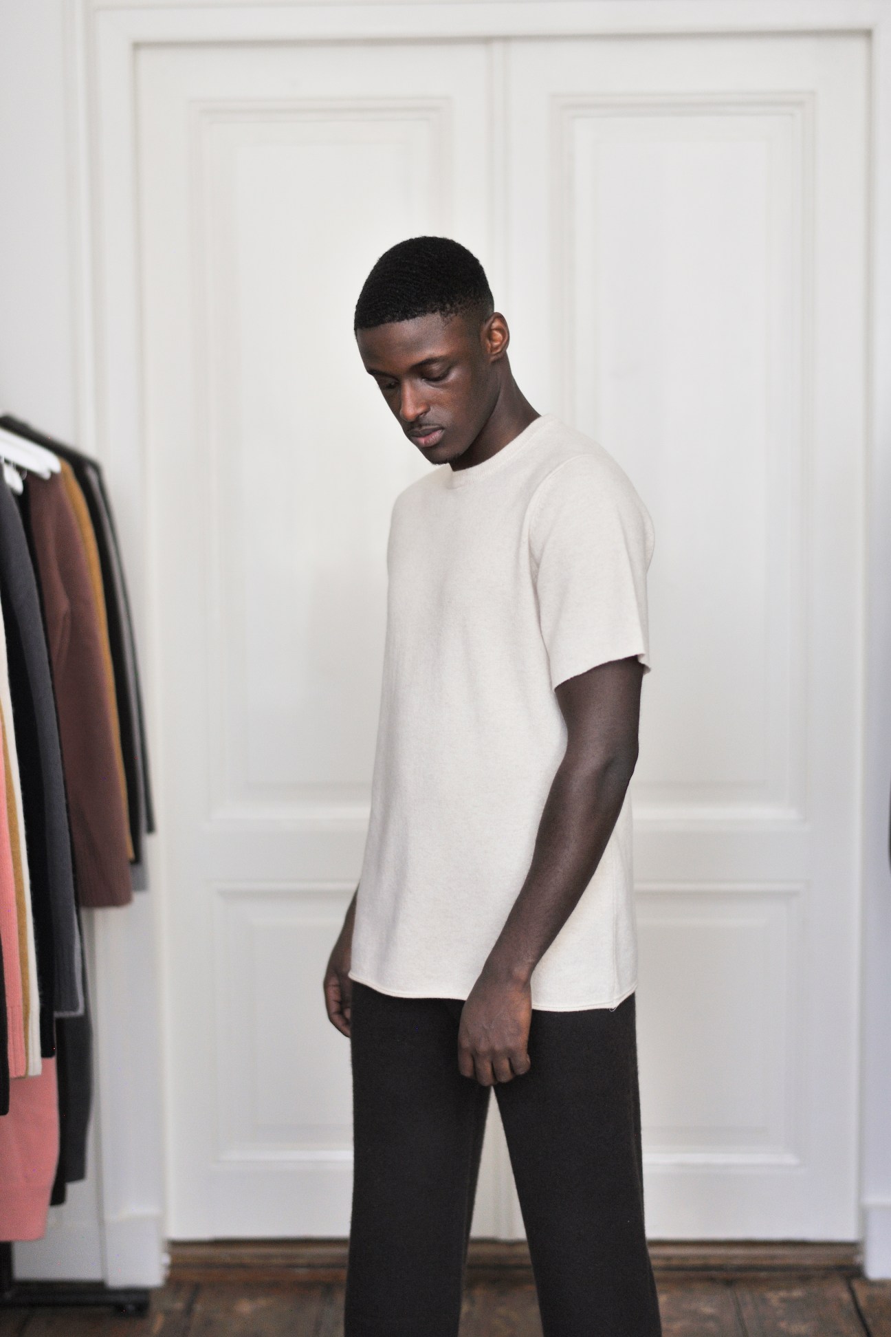 A person wearing a light-colored cashmere t-shirt and dark trousers, looking down in a minimal interior setting