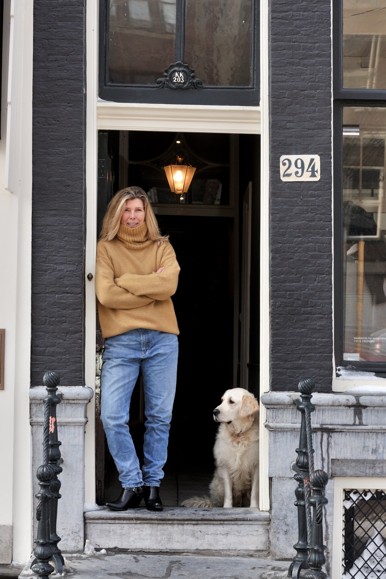 Woman in beige turtleneck sweater and jeans standing in doorway with golden retriever