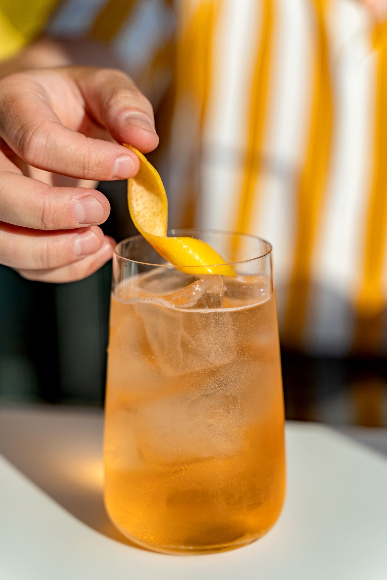 Hand adding orange peel garnish to an amber cocktail with ice in a glass