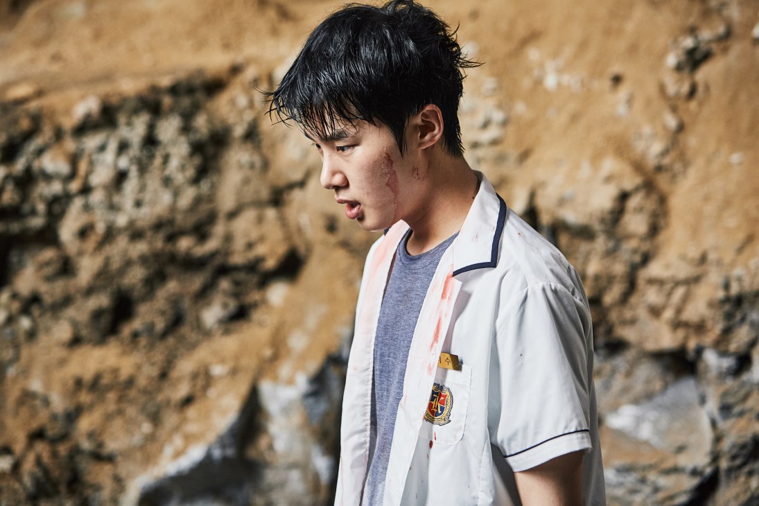 A young student wearing a white school uniform shirt stands against a textured brown wall.