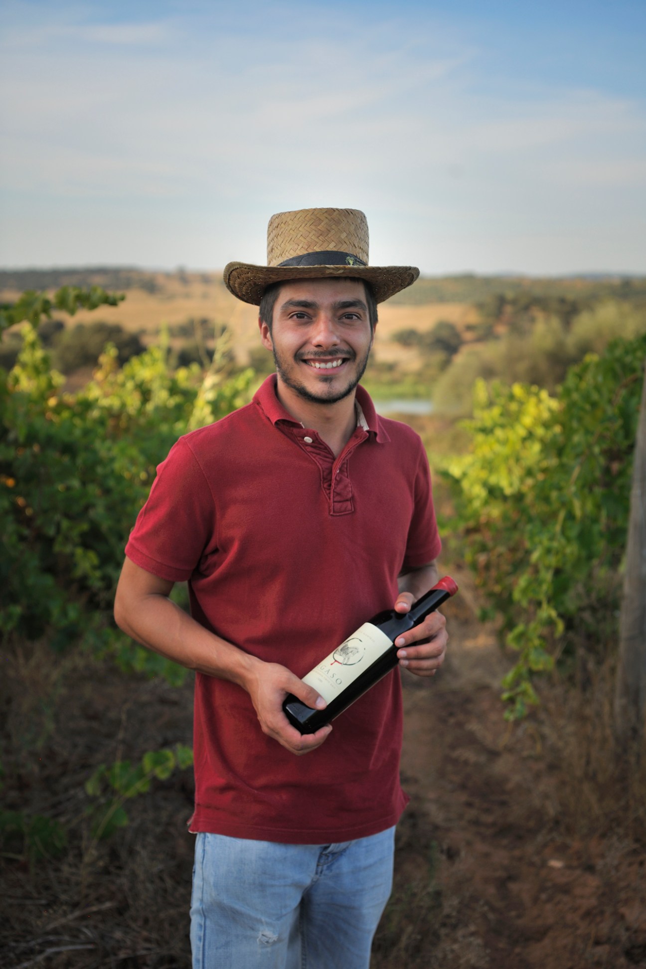 Young man in red polo shirt and straw hat holding wine bottle in vineyard