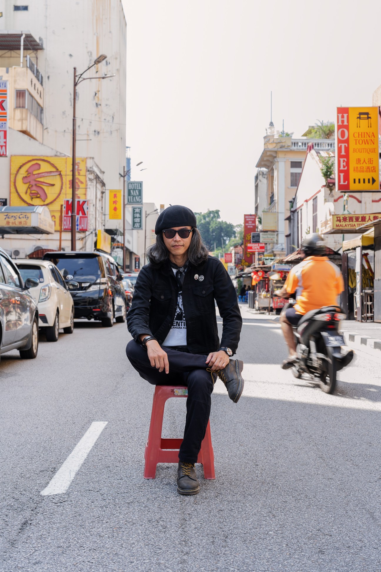 Fahmi Reza sits on a red stool in the middle of a street wearing sunglasses, black beret and dark clothing