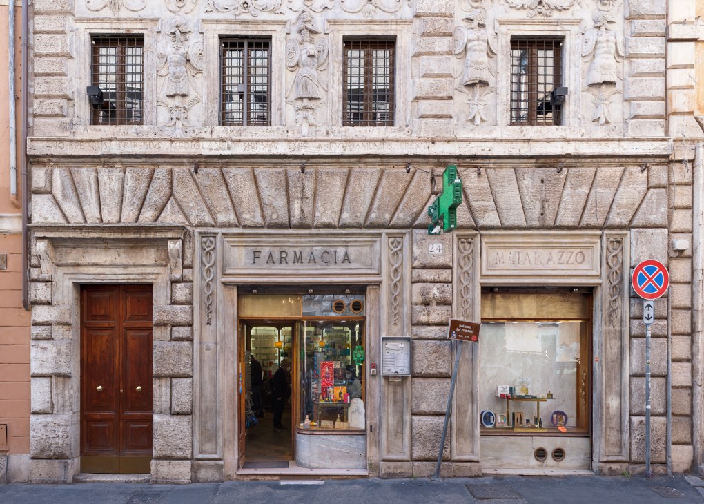 Historic Roman pharmacy storefront with ornate stone facade, green neon sign, and traditional wooden door.
