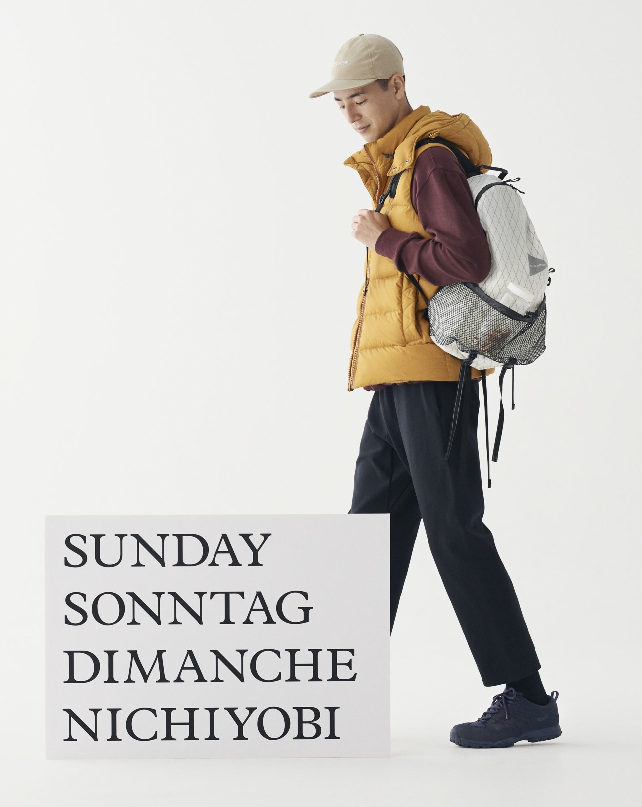 Model wearing mustard down vest, burgundy sweater, navy trousers, beige cap and backpack for outdoor Sunday look