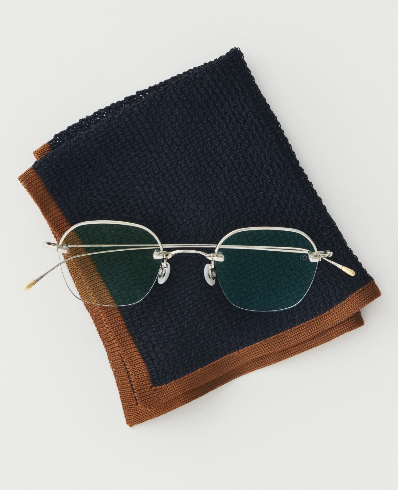 Rimless glasses with green-tinted lenses and gold temples on navy and brown textured fabric