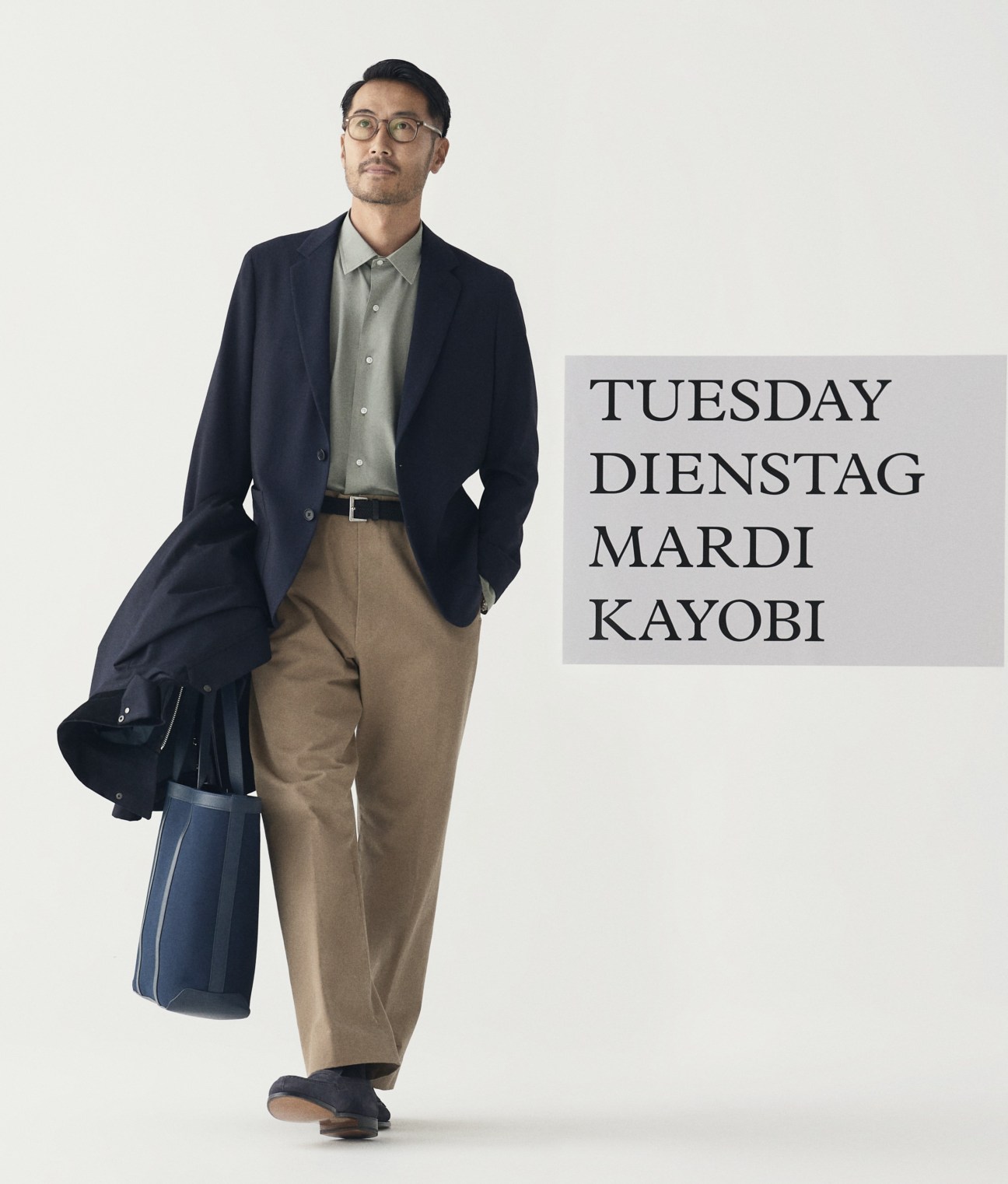 Model in beige coat, navy blazer, sage shirt, tan trousers with glasses, belt and bags