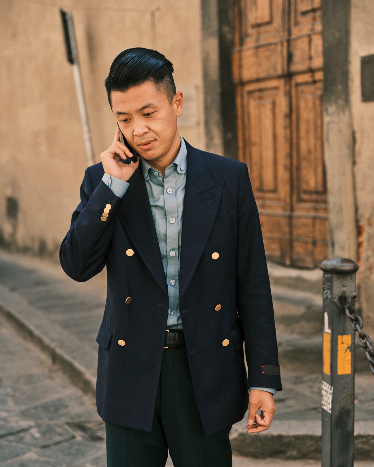 Man in navy double-breasted blazer talking on phone while walking on cobblestone street