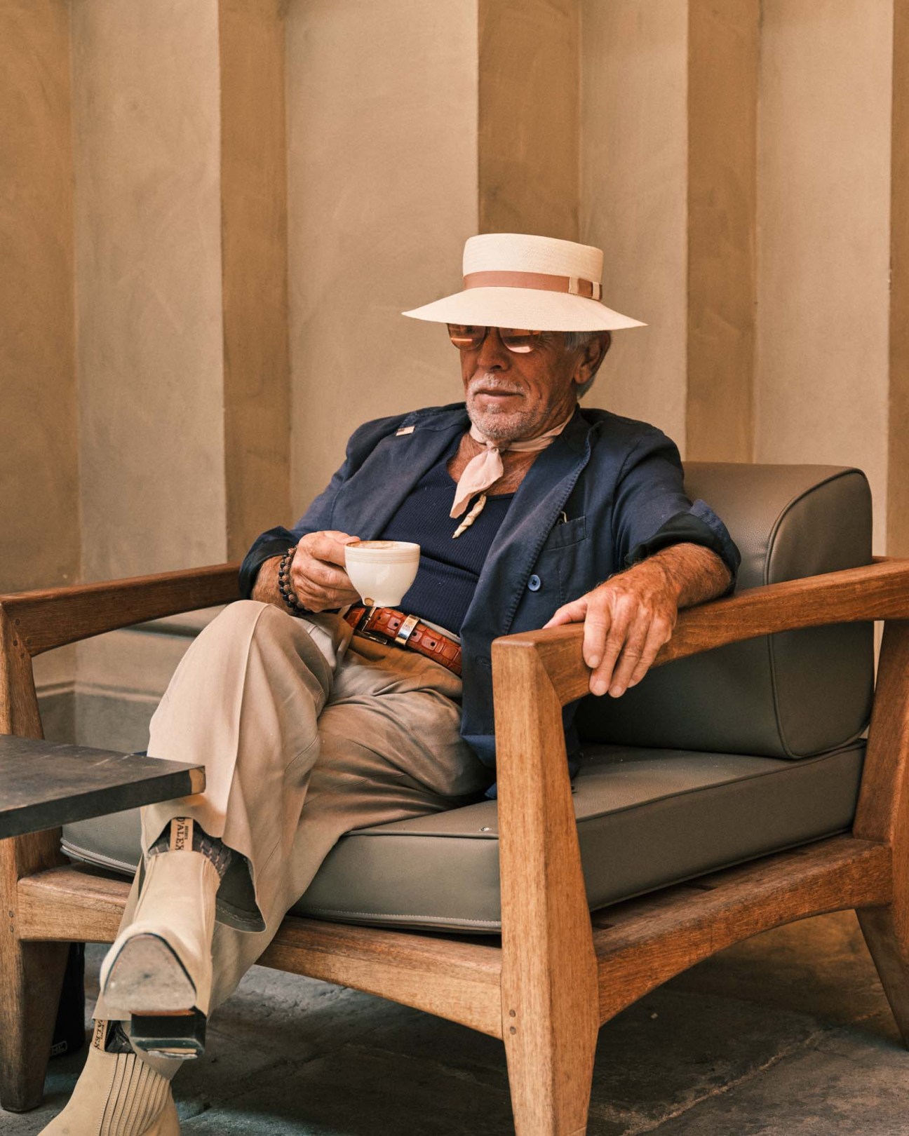 Older man in navy blazer and straw hat sits in wooden chair holding white coffee cup.