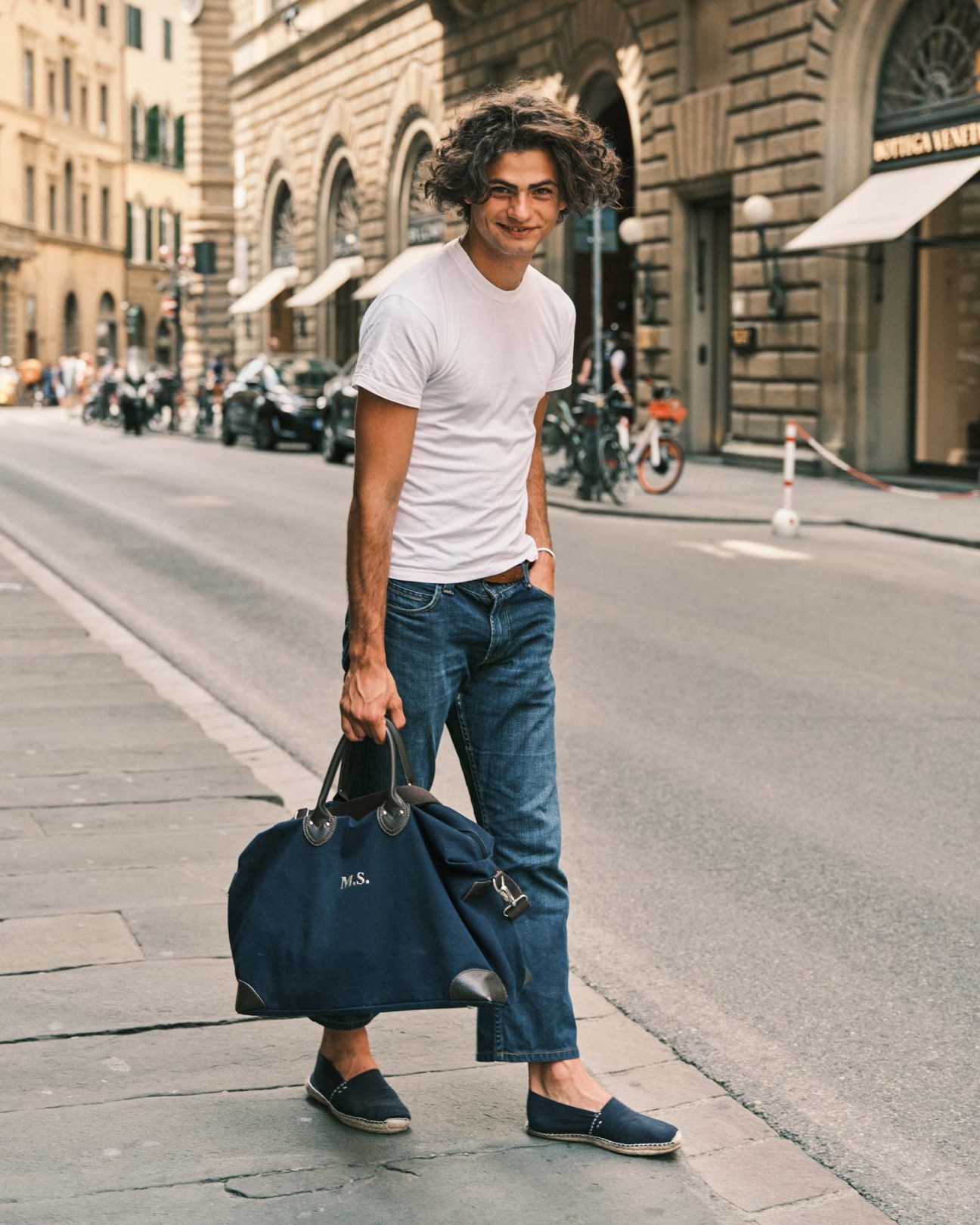 Man in white t-shirt and blue jeans standing on a Florence street holding a navy bag